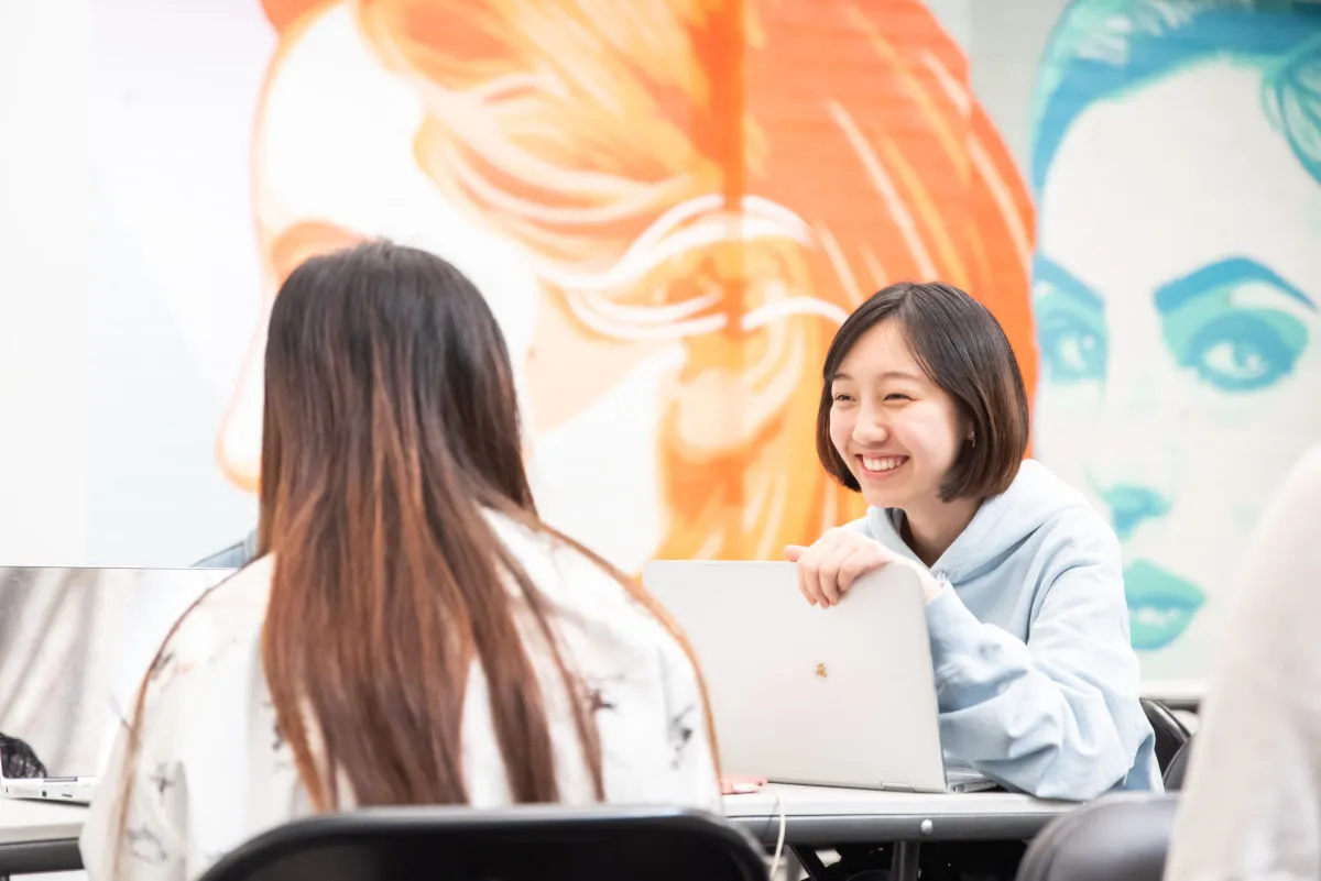 Two women chatting at a laptop in front of a mural