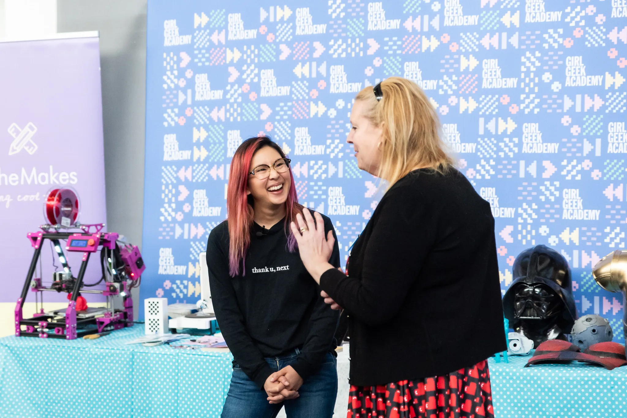 Two women chatting beside SheMakes banners and a 3D printer